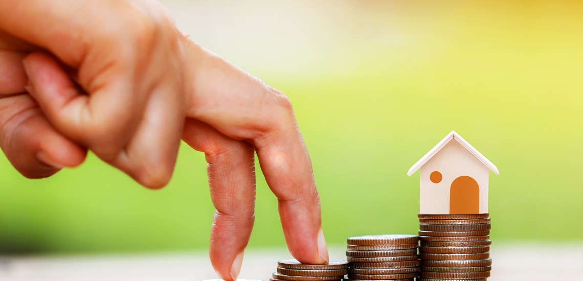 wooden house on piles of coins with hand walking up to the house